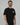 Black Red Shield Elite Mens Tee shown on a male model in a slight right-facing stance, highlighting the red Gracie Barra Red Shield emblem and red sleeve tag against a light gray background.
