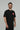 Black Red Shield Elite Mens Tee shown on a male model in a slight right-facing stance, highlighting the red Gracie Barra Red Shield emblem and red sleeve tag against a light gray background.